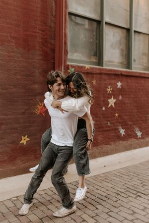 A joyful couple laughing as the man gives the woman a piggyback ride in front of a red brick wall with star murals.