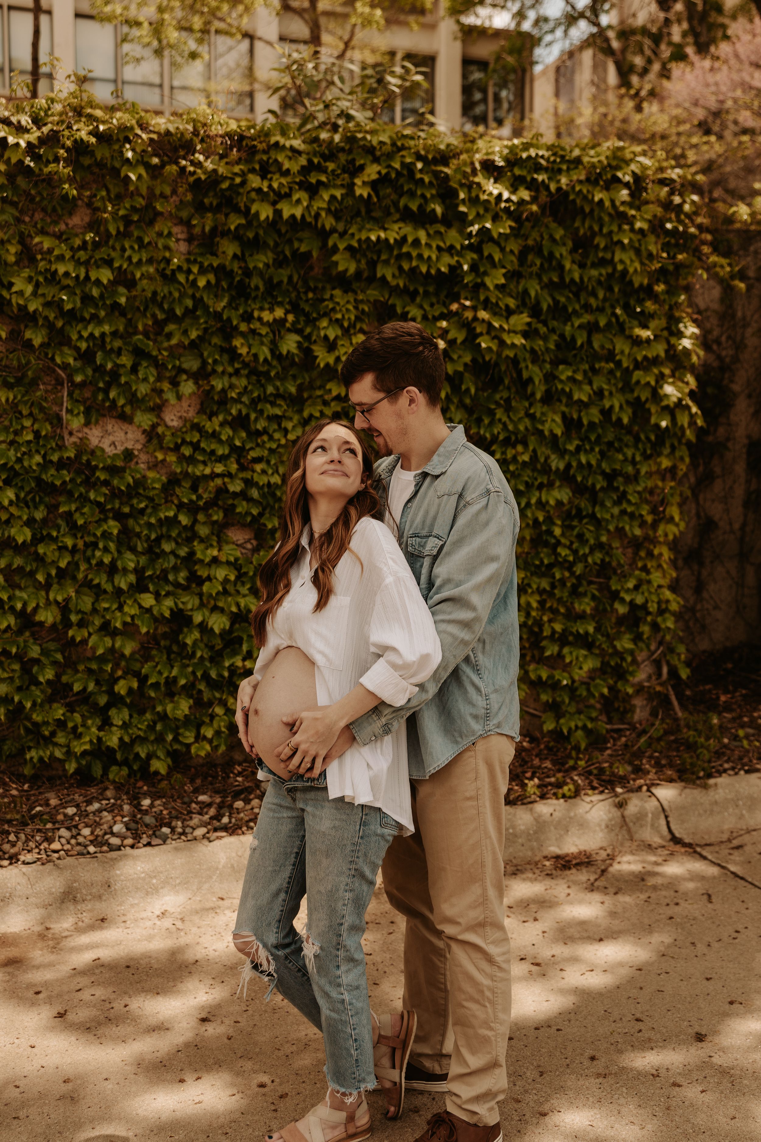 Pregnant couple embracing outdoors against ivy-covered wall, woman in white top and jeans, man in green shirt and khakis.