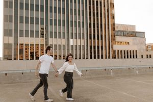 A young couple holds hands and walks across an urban rooftop parking lot with a modern glass building in the background.