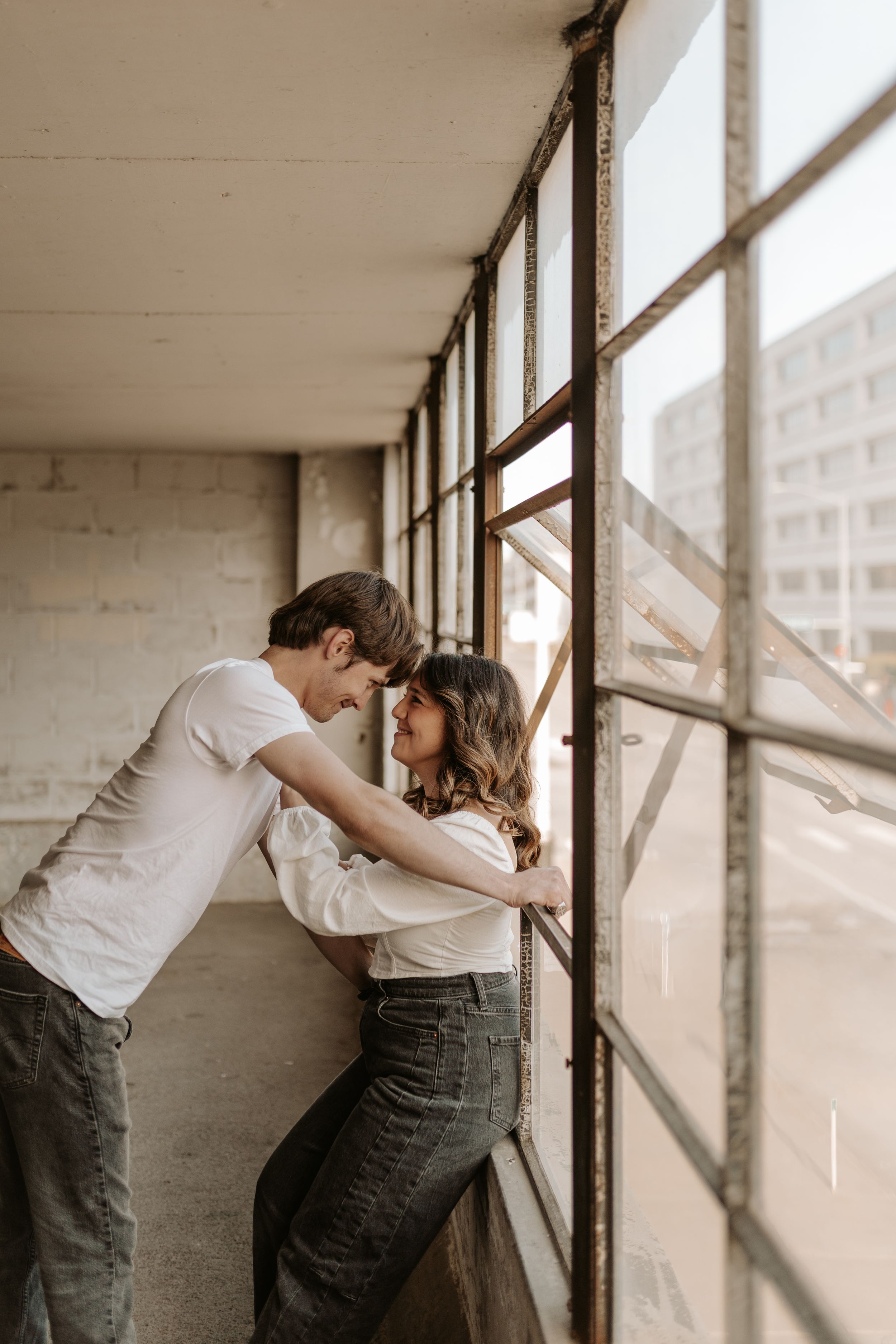 A young couple shares a romantic moment leaning toward each other by large industrial windows in an urban building.