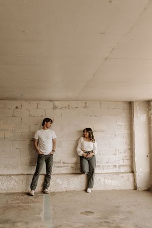 A couple in white tops and gray jeans lean against a white brick wall in an unfinished concrete space.