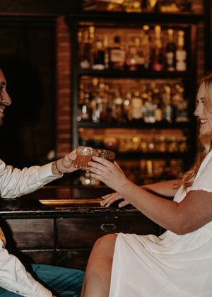 Couple in white attire toasting cocktails at a dimly lit bar with bottle-lined shelves in the background.