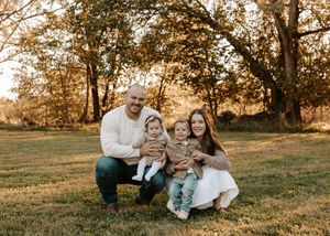Smiling family of four with two young children posing together on a grassy lawn surrounded by autumn trees in warm golden light.