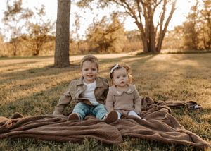 Two young children sit side by side on a brown knit blanket in a sunny grassy park surrounded by trees in warm golden light.