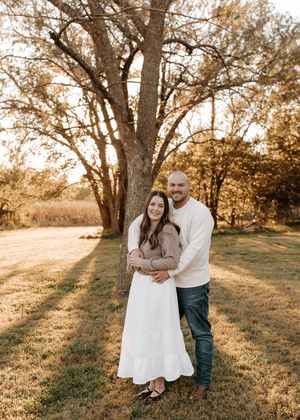 Smiling couple embracing under a large tree in a golden grassy field at sunset, she in a white skirt and he in a cream sweater.