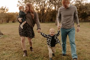 Smiling family of four walks hand in hand across a grassy field at sunset, with a mother carrying a baby and a father leading a toddler.