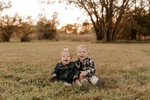 Two toddlers in matching denim and plaid outfits sit side by side on a grassy field with tall trees in the background at golden hour.