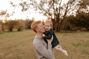 A man in a gray sweater kisses the cheek of a smiling baby girl in a green dress while holding her outdoors in a grassy field at sunset.