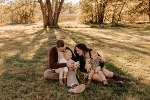 A family of four sits together on a sunlit grassy field with trees behind them, as two toddlers snuggle in their parents' laps.