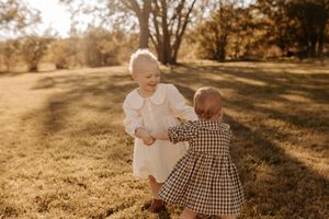 Two toddlers in matching vintage outfits hold hands and play in a sunny autumn field with golden grass and trees in the background.