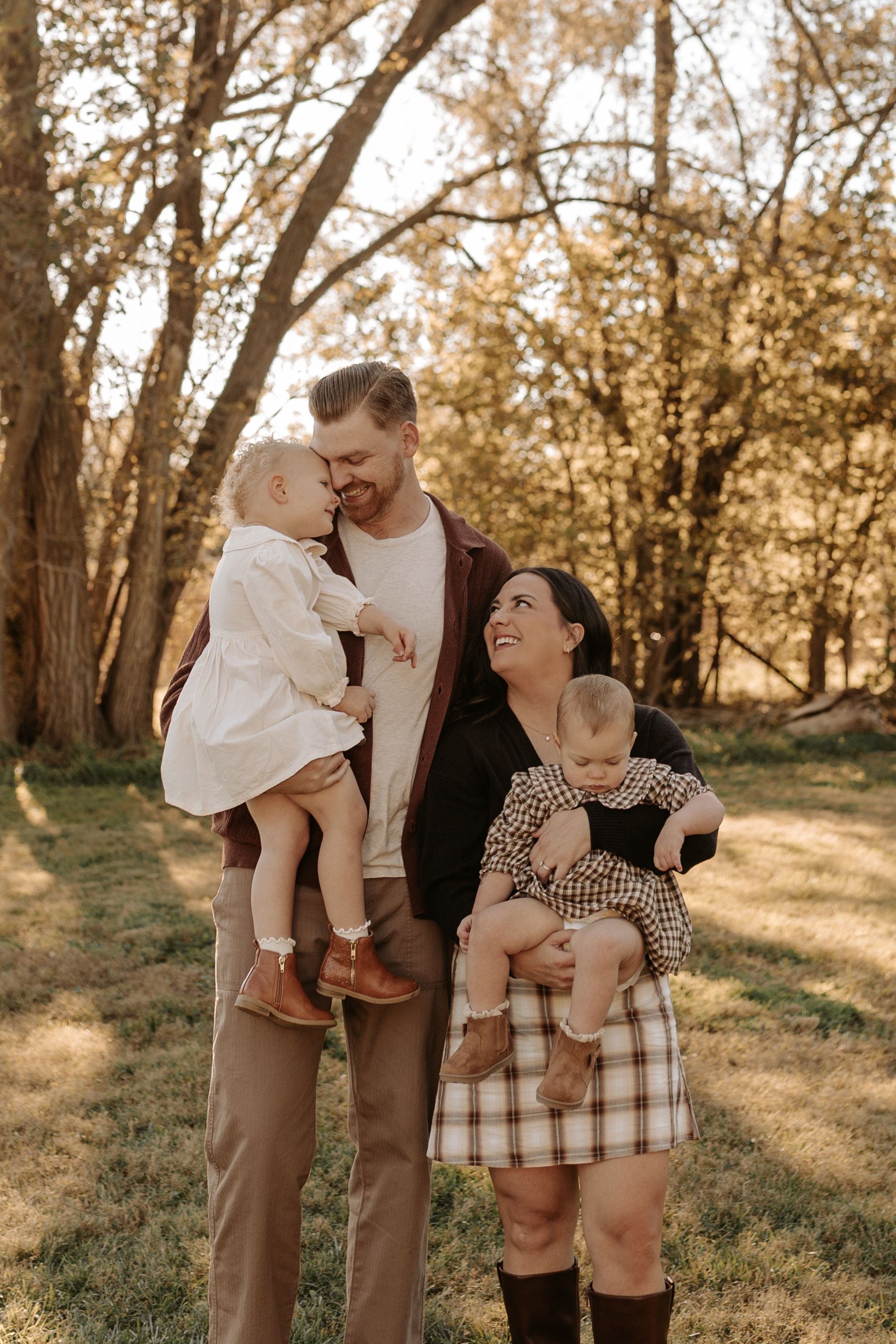 A smiling couple holds two toddlers in an outdoor autumn family portrait, standing among trees in warm golden sunlight.