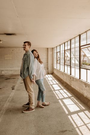 A couple stands back-to-back in a sunlit industrial hallway, smiling, dressed in casual earth-toned outfits.