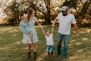 Family of four enjoying a sunny outdoor walk, parents swinging toddler boy between them on lush green grass.