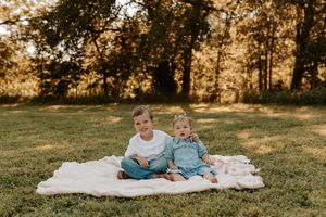 Two young siblings sit together on a blanket in a sunny green park, smiling for a family photo.