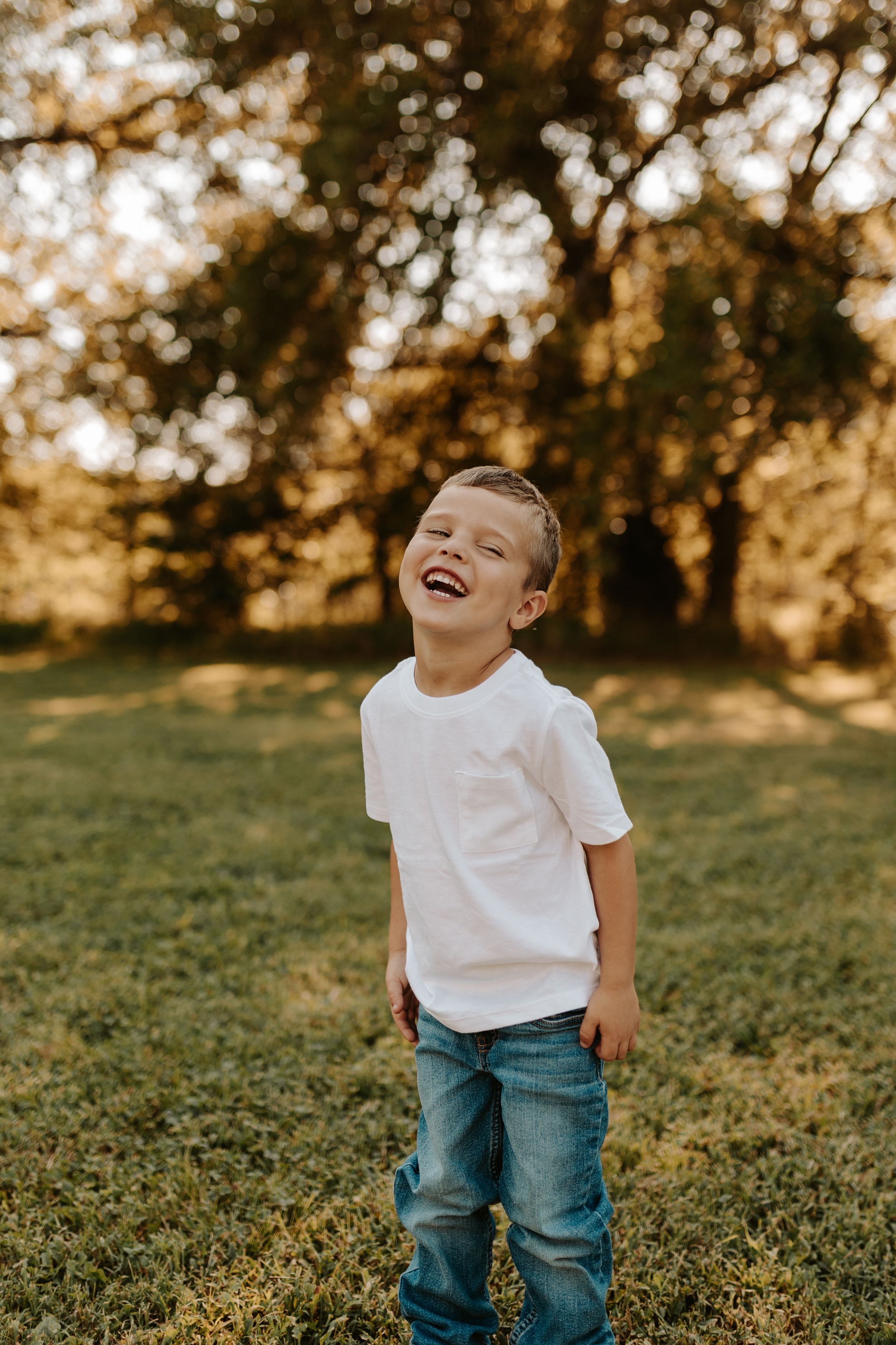 Happy young boy laughing outdoors in white t-shirt and jeans, standing on green grass with trees in golden sunlight.