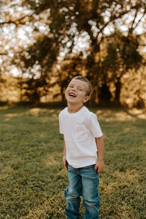 Happy young boy laughing outdoors in white t-shirt and jeans, standing on green grass with trees in golden sunlight.
