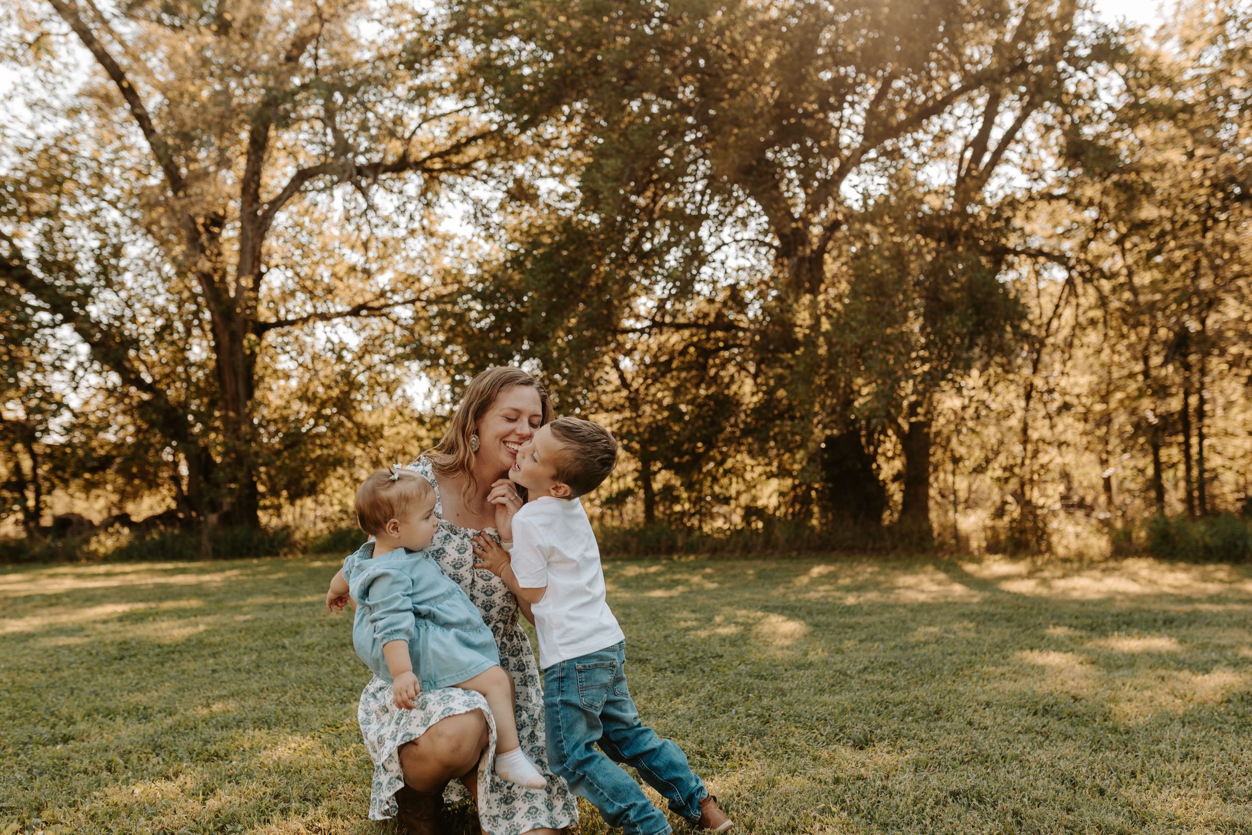 A mother sits outdoors in golden sunlight, lovingly embracing her two young children in a park.