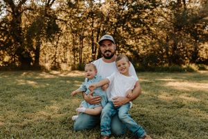 A bearded father in a white tee and cap sits on grass holding two young toddlers in blue outfits outdoors.