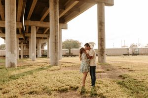 Couple sharing a romantic kiss under a concrete highway overpass on a sunny day in an urban outdoor setting.