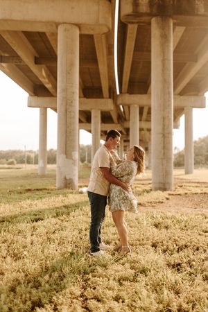 Couple embracing romantically under a concrete bridge overpass in a golden field during sunset.