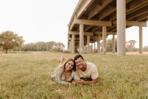 Couple lying in green grass smiling, with large concrete bridge pillars and trees in the background.
