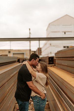 Couple sharing a romantic kiss between rustic steel beams at an industrial outdoor location on a cloudy day.