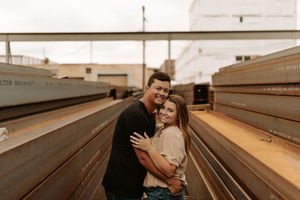 Couple embracing and smiling between stacked steel beams at an industrial outdoor location during golden hour.