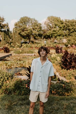 Young man in light blue shirt and beige shorts standing in a lush garden with colorful plants and trees.