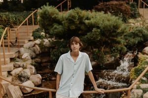 Young man in light blue short-sleeve shirt stands on wooden bridge by waterfall in lush garden.