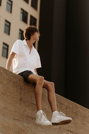 Young man in white shirt, black shorts, and white sneakers sitting on a concrete ledge outside a building.