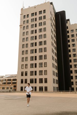 A woman in white shirt and black shorts stands in an open plaza before a tall beige multi-story building.
