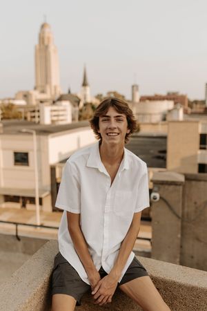 Smiling teen boy in white short-sleeve shirt sitting on rooftop ledge with city skyline behind him.