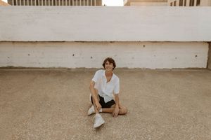 Young person in white shirt and dark shorts sitting relaxed on ground against white wall outdoors.