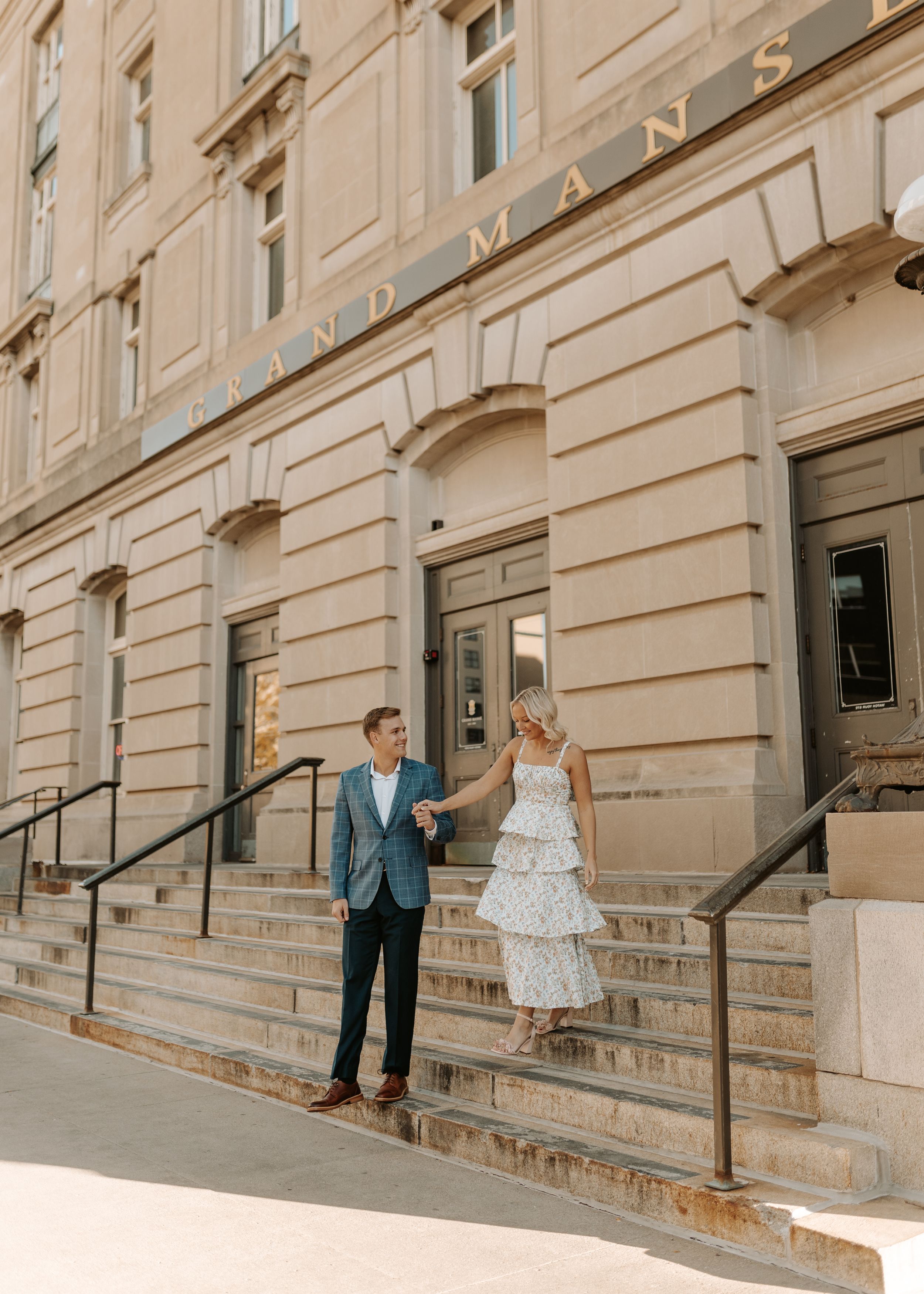 Couple holding hands on steps outside Grand Mansion building, man in blue suit and woman in white tiered dress.