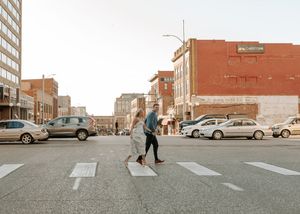 Couple crossing a busy urban street crosswalk in a downtown city area with brick buildings and parked cars.
