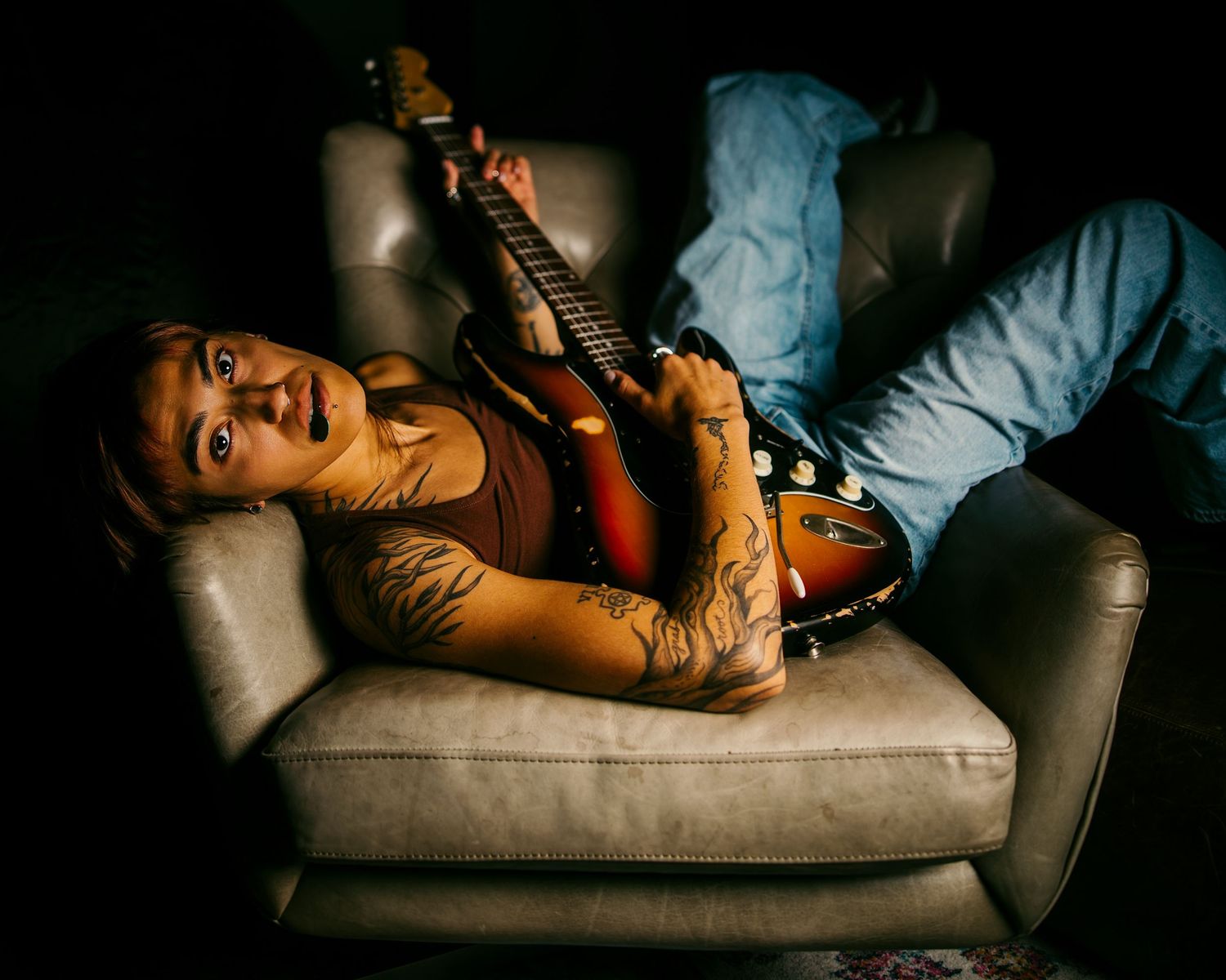 Tattooed woman lying on gray sofa holding a sunburst electric guitar in dim light.