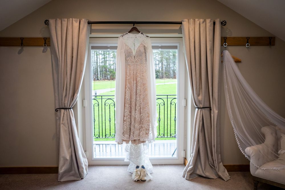 Elegant lace wedding dress hanging in a window with soft drapes and bridal shoes below in a bright room.