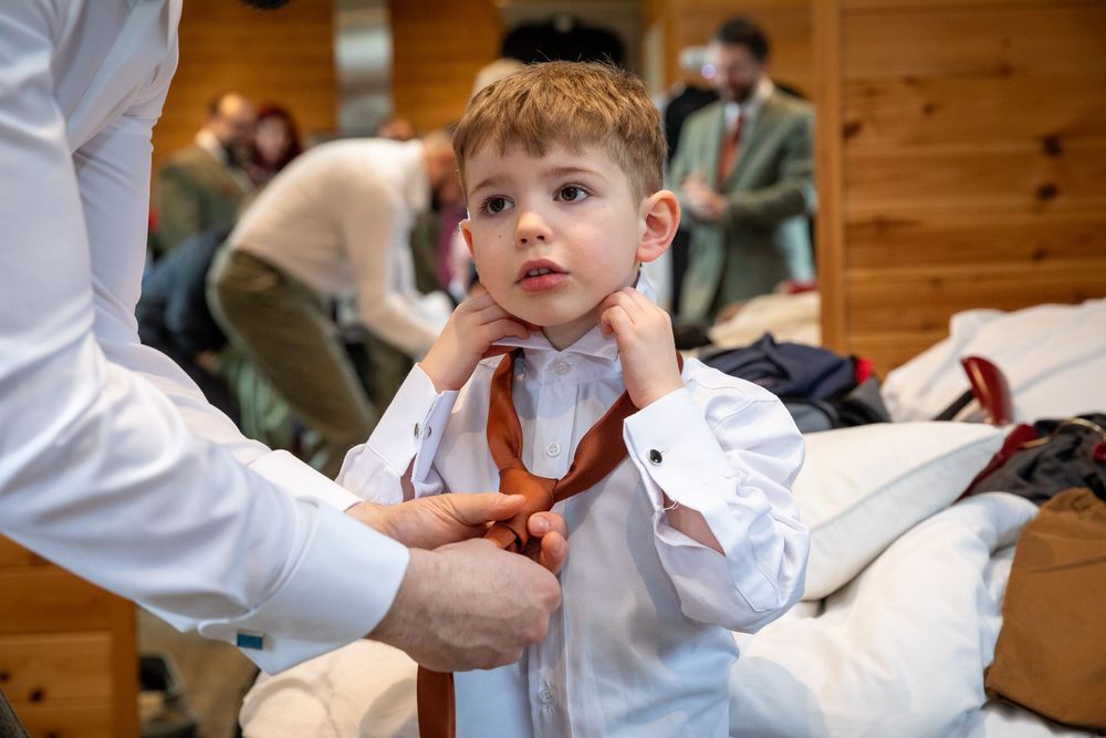 Young boy in white shirt getting his brown tie adjusted by an adult during wedding preparations.