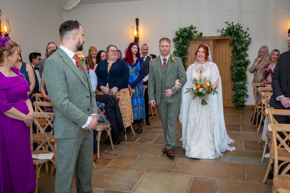 Bride walks down the aisle with her father at a rustic wedding ceremony, guests watching in colorful attire.