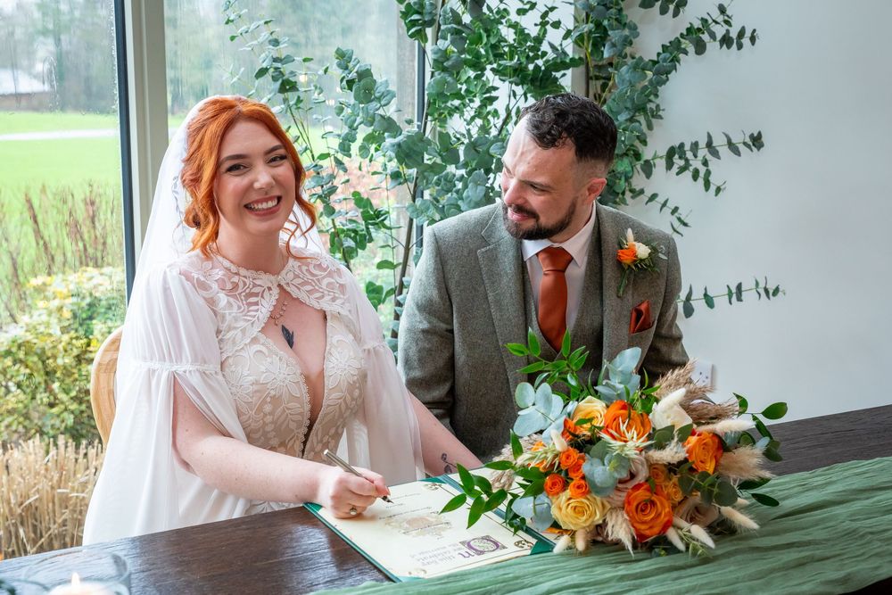 Bride signing wedding register smiling beside groom in grey suit with orange tie and boutonniere, orange bouquet on table.