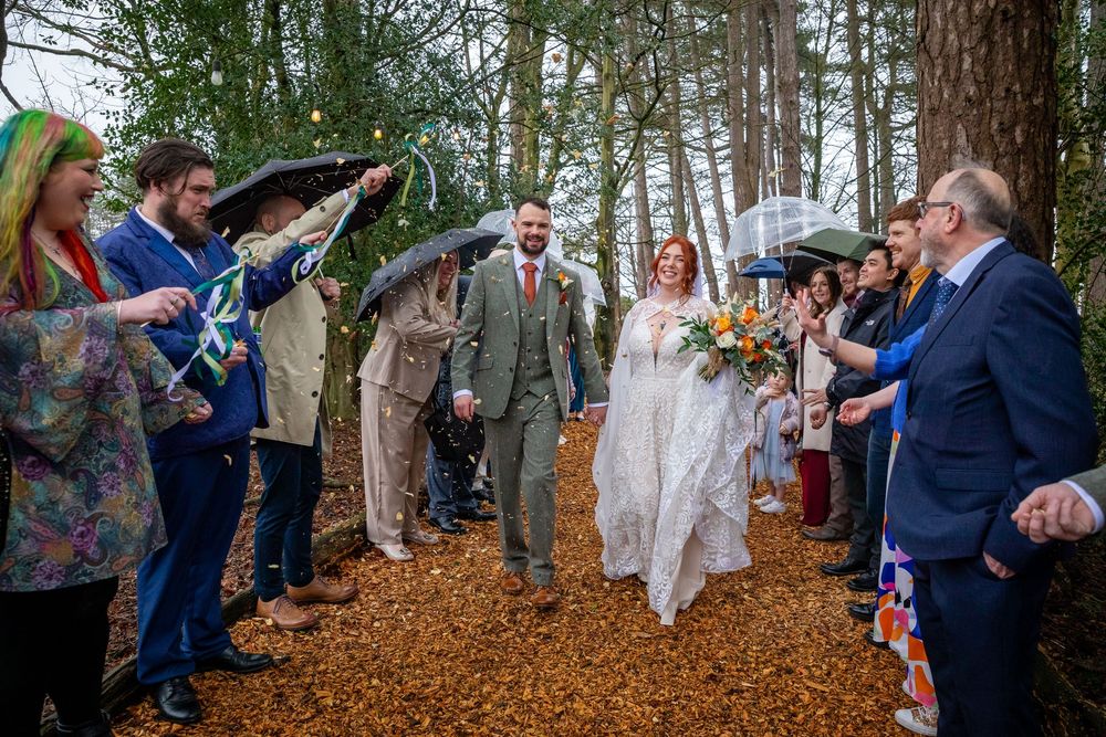 Bride and groom walk through woodland aisle lined with guests holding umbrellas on a rainy autumn wedding day.