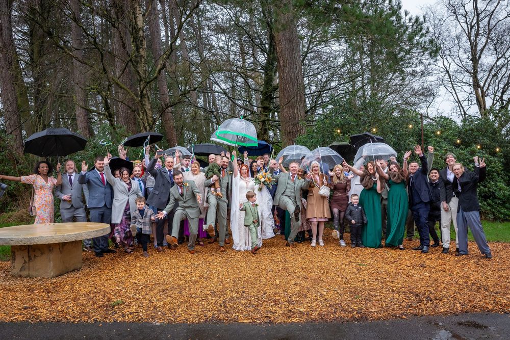 Wedding party celebrating outdoors in the rain, holding umbrellas on a leaf-covered ground surrounded by trees.