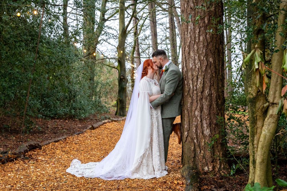 A bride and groom share a romantic kiss among tall trees on a forest path covered in autumn leaves.