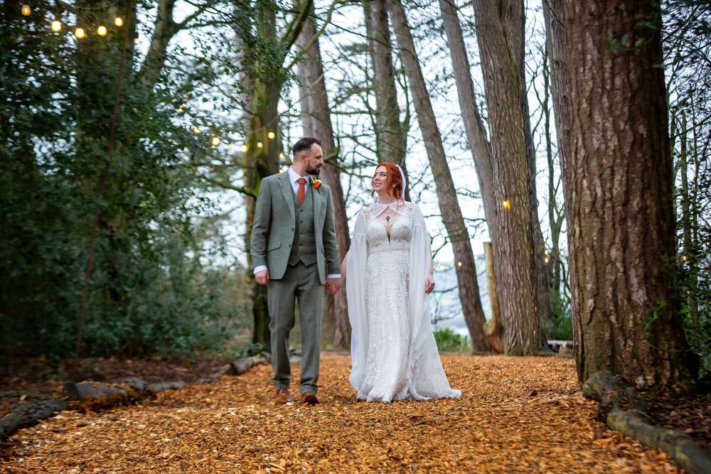 Bride and groom walking hand in hand through autumn woodland with fairy lights among tall trees.