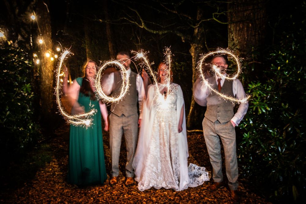 Wedding party writing LOVE with sparklers at night in a forest setting, bride in white gown with guests in formal attire.