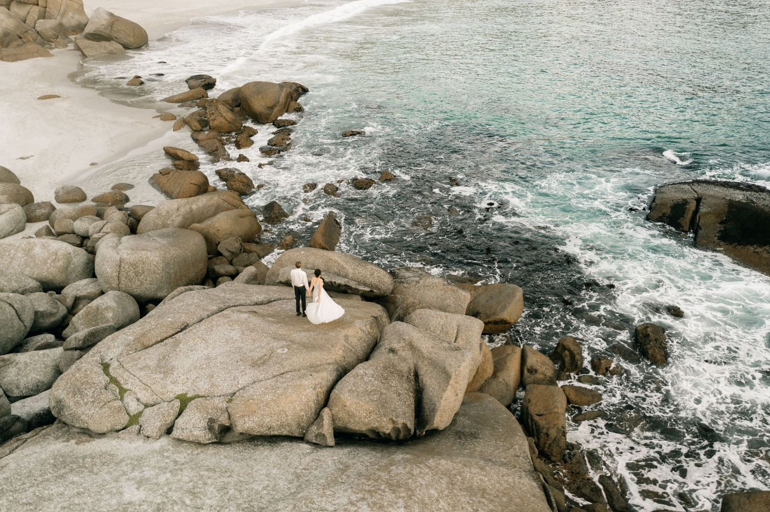 Aerial view of a wedding couple standing on rocky coastal boulders with turquoise ocean waves crashing around them.