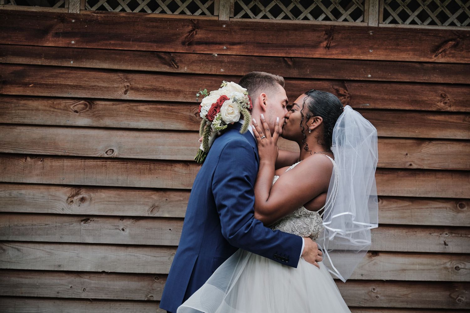 A bride in a white gown and groom in navy suit share a romantic kiss against rustic wooden barn siding.
