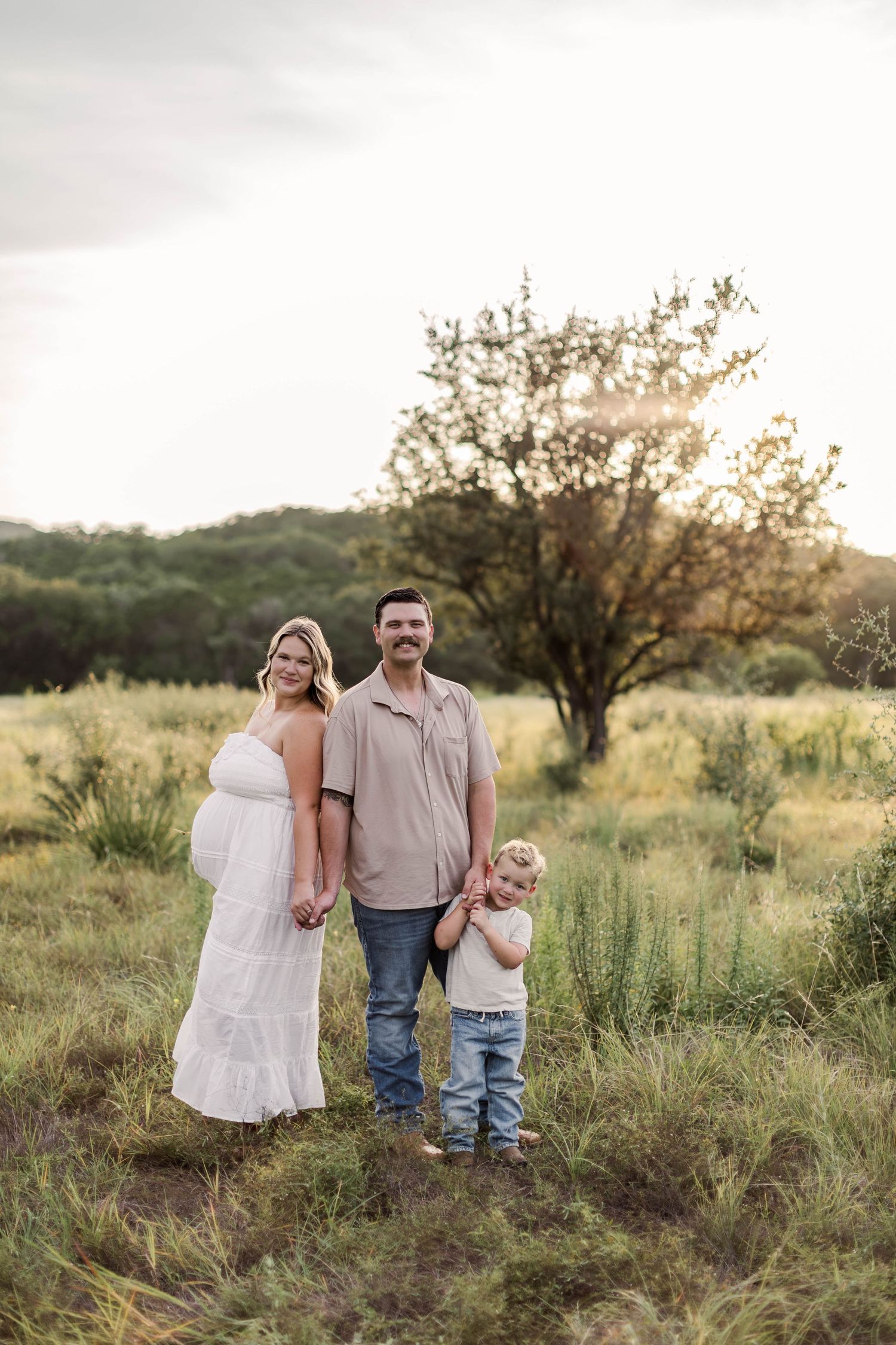 Pregnant woman in white dress with husband and toddler in sunny green meadow during golden hour maternity photo session.