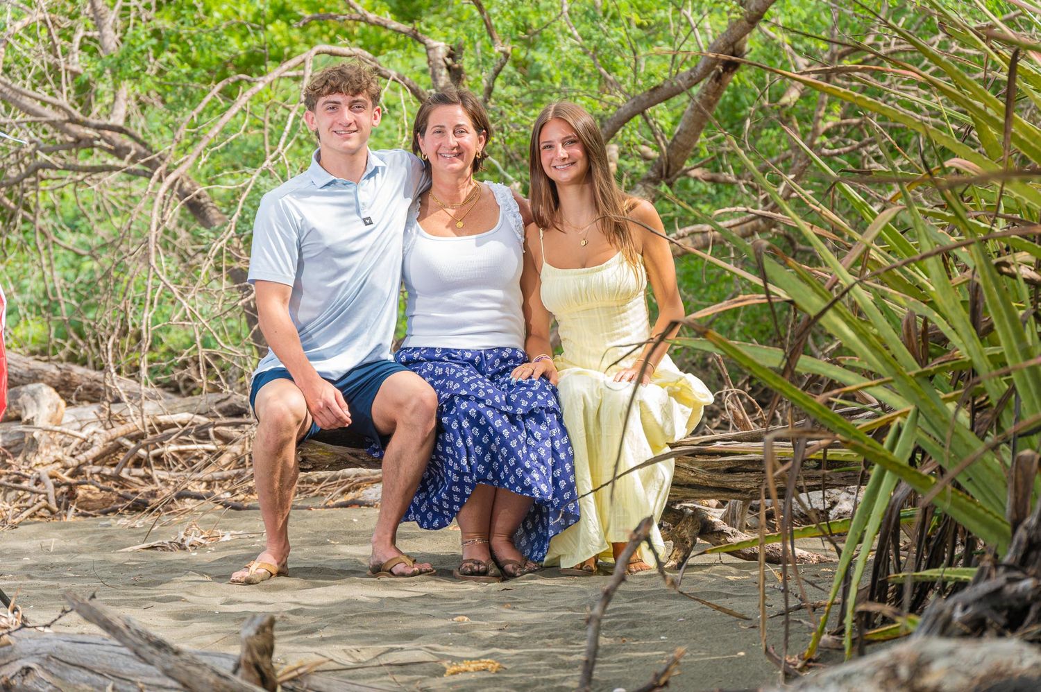 Three people smiling together on a wooden bridge surrounded by tropical mangrove trees in a lush outdoor setting.