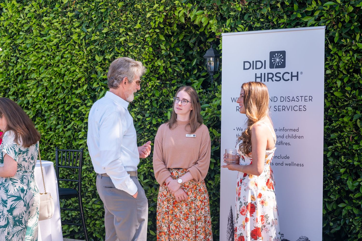 Three people networking at a Didi Hirsch Mental Health Services outdoor event near a branded banner.